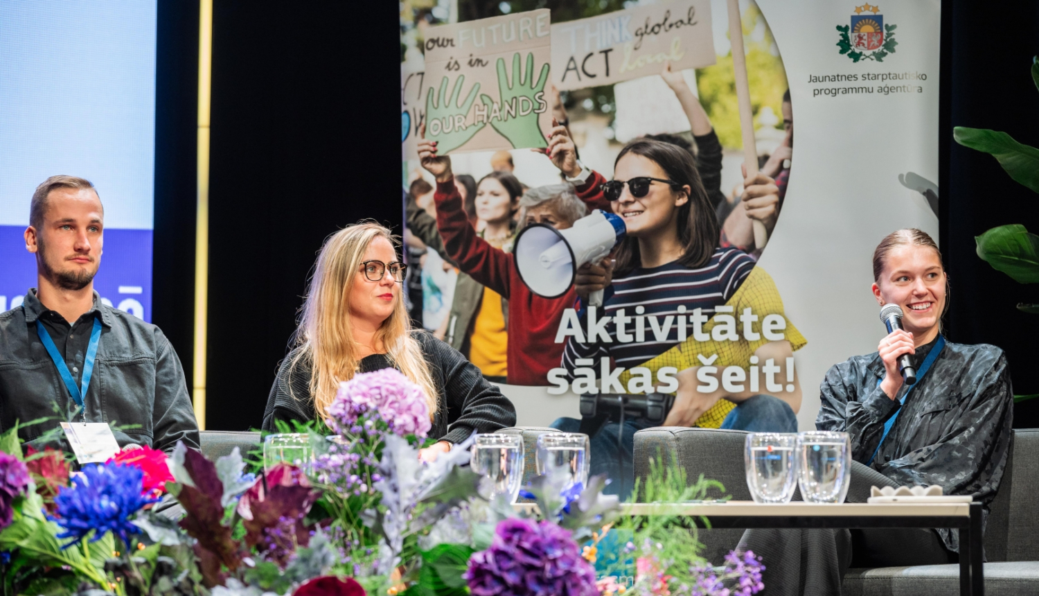 Viens jauns vīrietis un divas jaunas sievietes sēž uz skatuves un runā, priekšplānā ziedu pušķis, aizmugurē plakāts, uz kura attēloti jaunieši un uzraksts "Aktivitāte sākas šeit!"