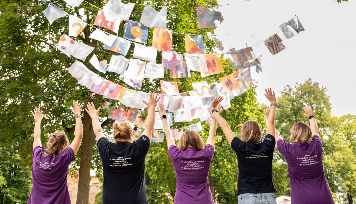 Piecas meitenes redzamas no mugupuses, viņas ir pacēlušas rokas uz augšu, augšā kokos iekārtas virtenes