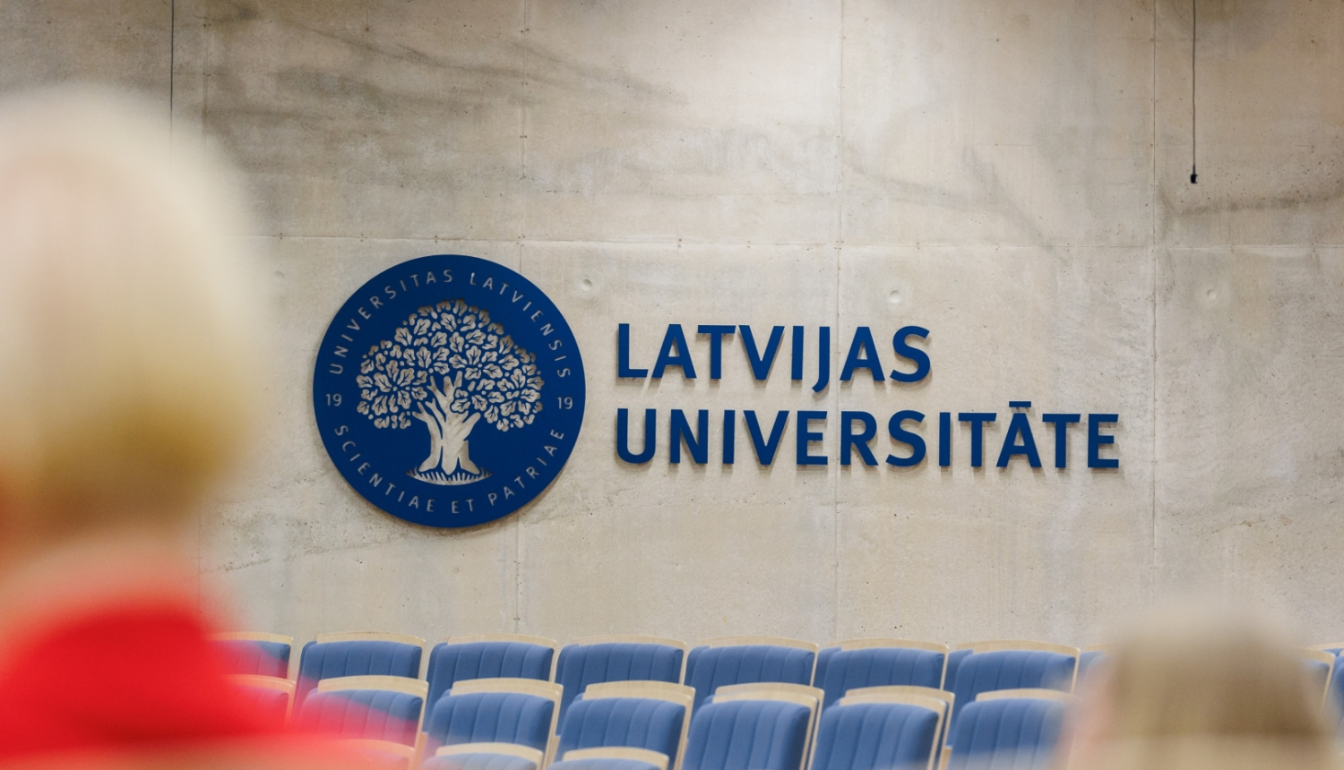 Latvijas Universitātes auditorija ar ziliem krēsliem, uz sienas liels zils LU logotips - ozols zilā aplī un uzraksts Latvijas Universitāte