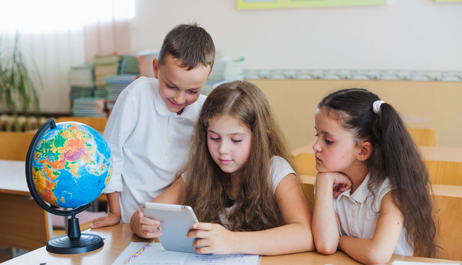 Trīs bērni ar planšeti sēž pie galda, uz galda globuss