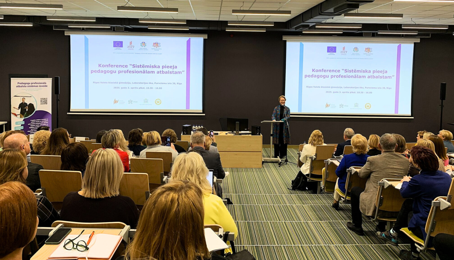 Cilvēku grupa sēž un klausās konferenci. Konferenci atklāj izglītbas un zinātnes ministre Dace Melbārde.