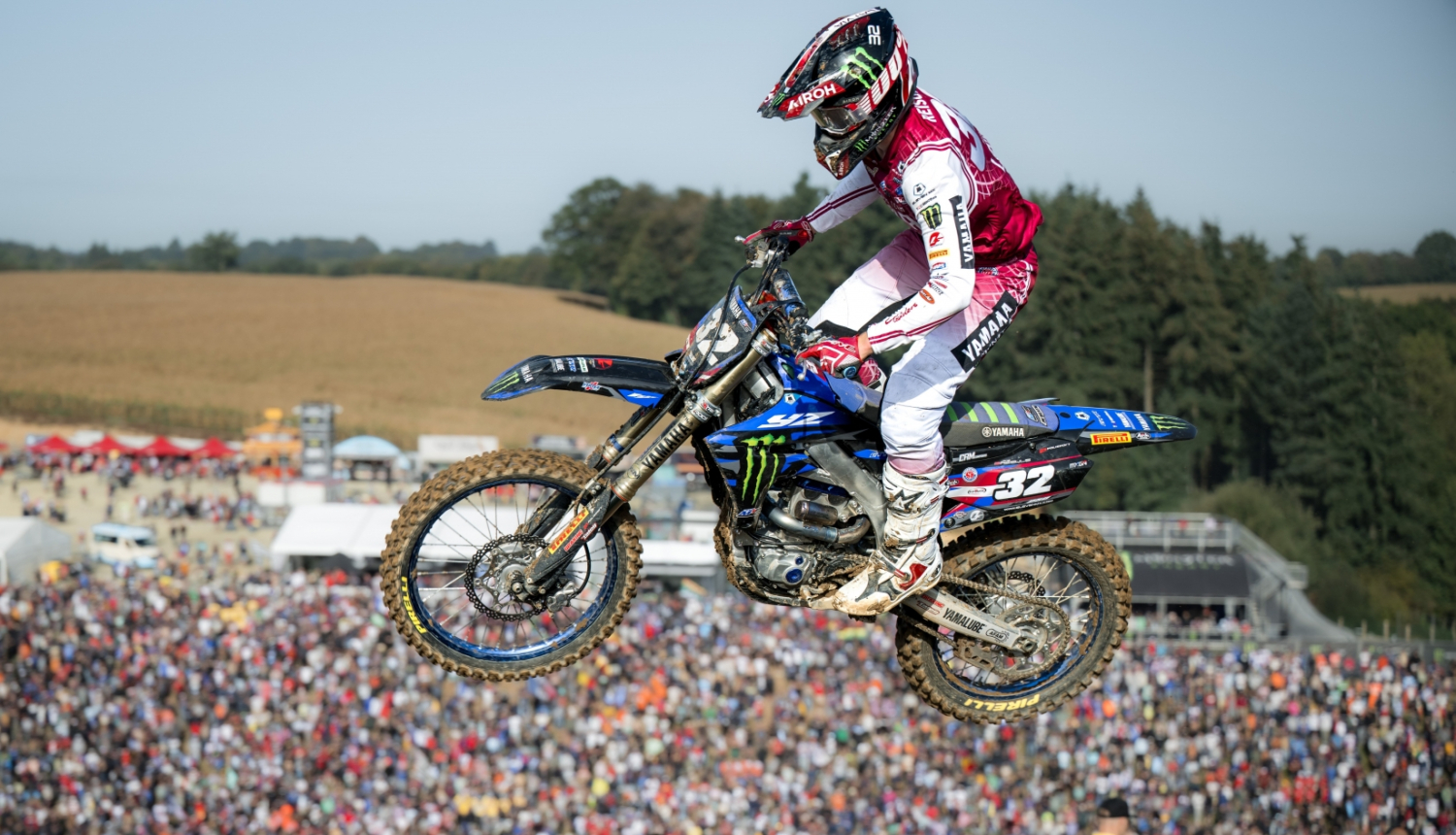 Motokrosa sportists uz motocikla ir palēcies un kadrā notverts gaisā, zem viņa tālumā ir daudz skatītāju 