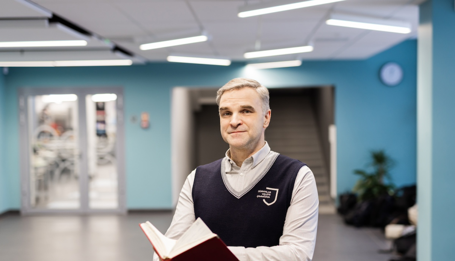 Attēlā redzams skolotājs Jānis no Jūrmalas Valsts ģimnāzijas, kurš rokās tur grāmatu