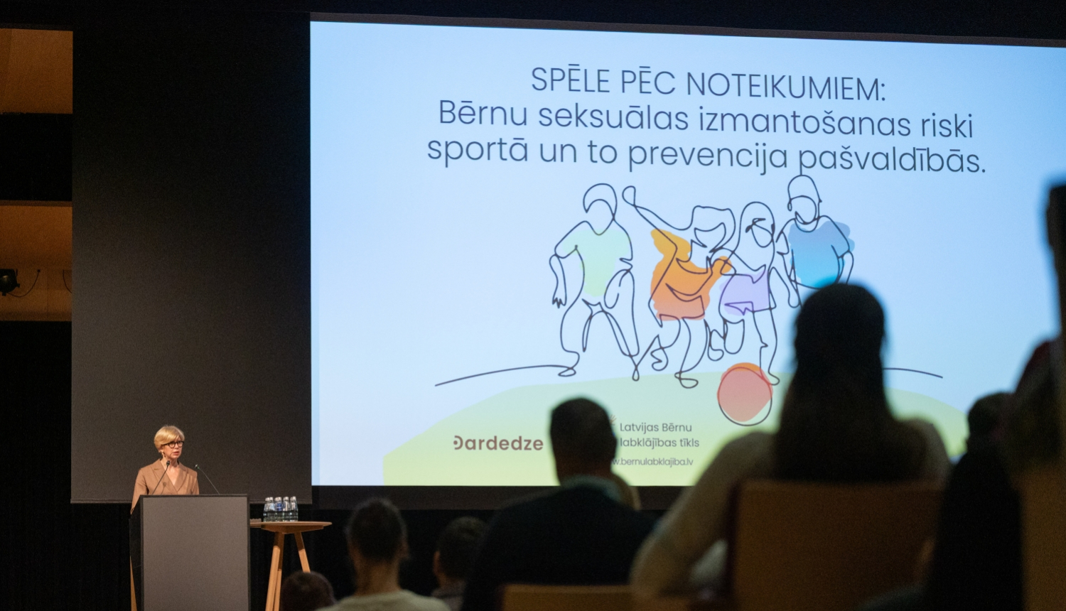 Izglītības un zinātnes ministre Anda Čakša konferencē uz skatuves, aiz viņas liels ekrāns ar bērnu ilustratīvu zīmējumu