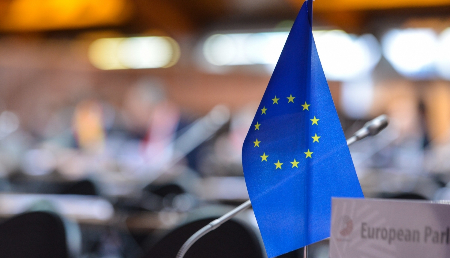 Priekšplānā Eiropas Savienības karodziņš, aizmugurē konferences telpas sēdvietas