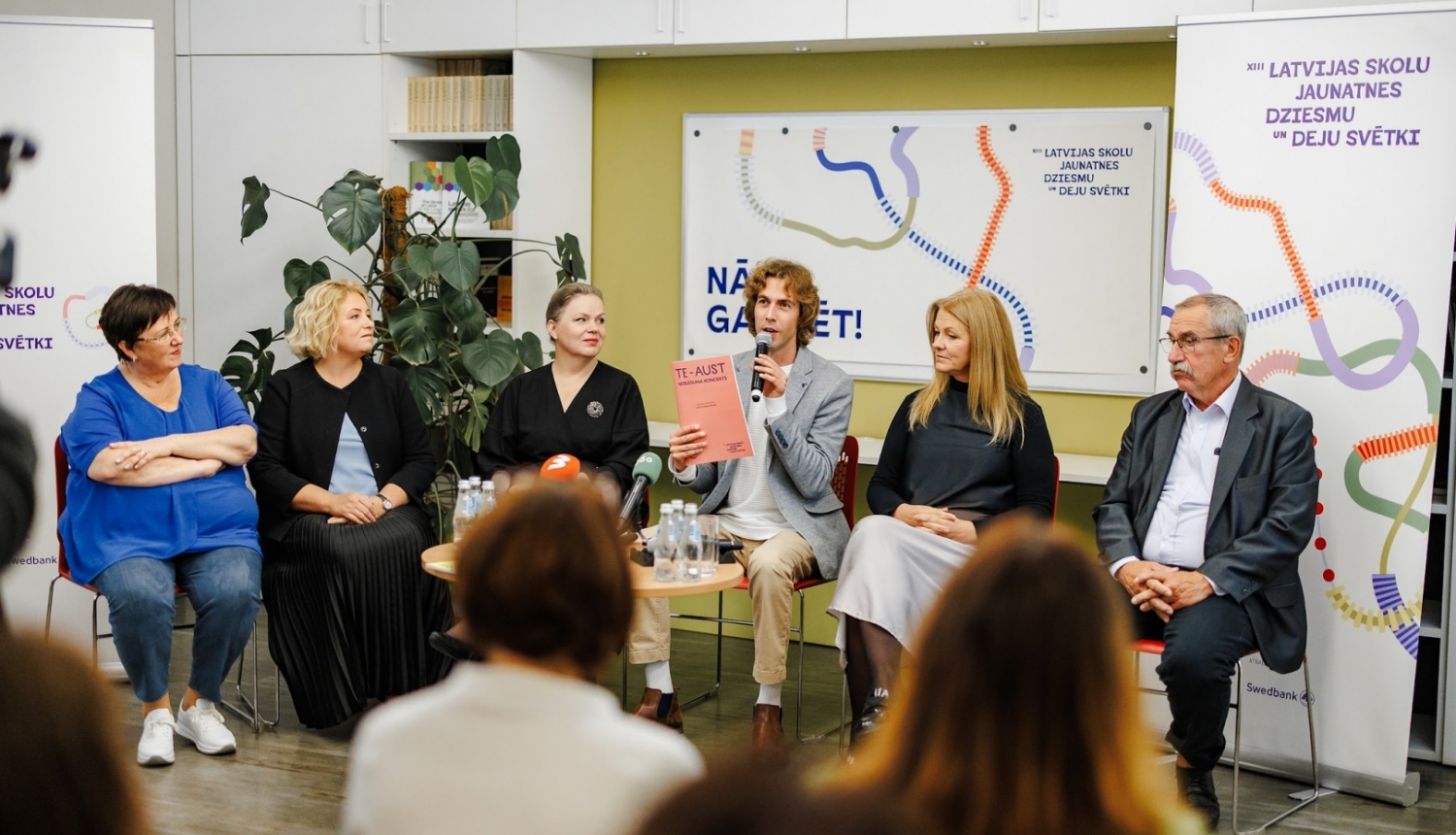 XIII Latvijas Skolu jaunatnes dziesmu un deju svētku preses konference. Skats no zāles, priekšā sēž seši cilvēki un stāsta par pasākuma norisi.