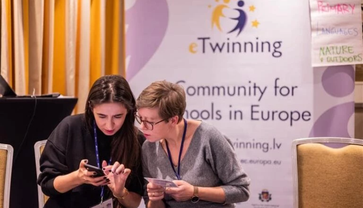 eTwinning baneris, priekšā tam sēž divas sievietes un pēta telefonu