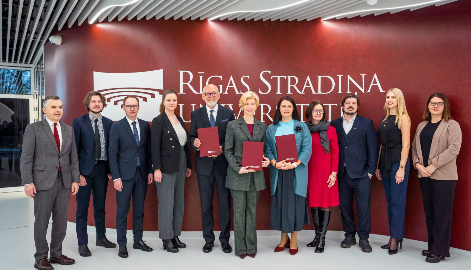 RSU un IZM pārstāvju kopbilde pie sienas, kur redzams Rīgas Stradiņu universitātes logo.
