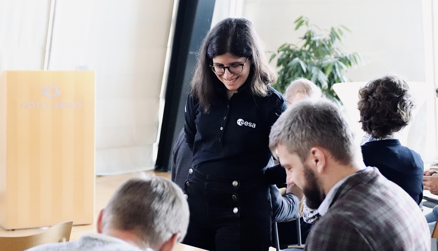 Smaidīga meitene stāv blakus galdam, kur sēž divi smaidīgi vīrieši. Notiek mācību process.