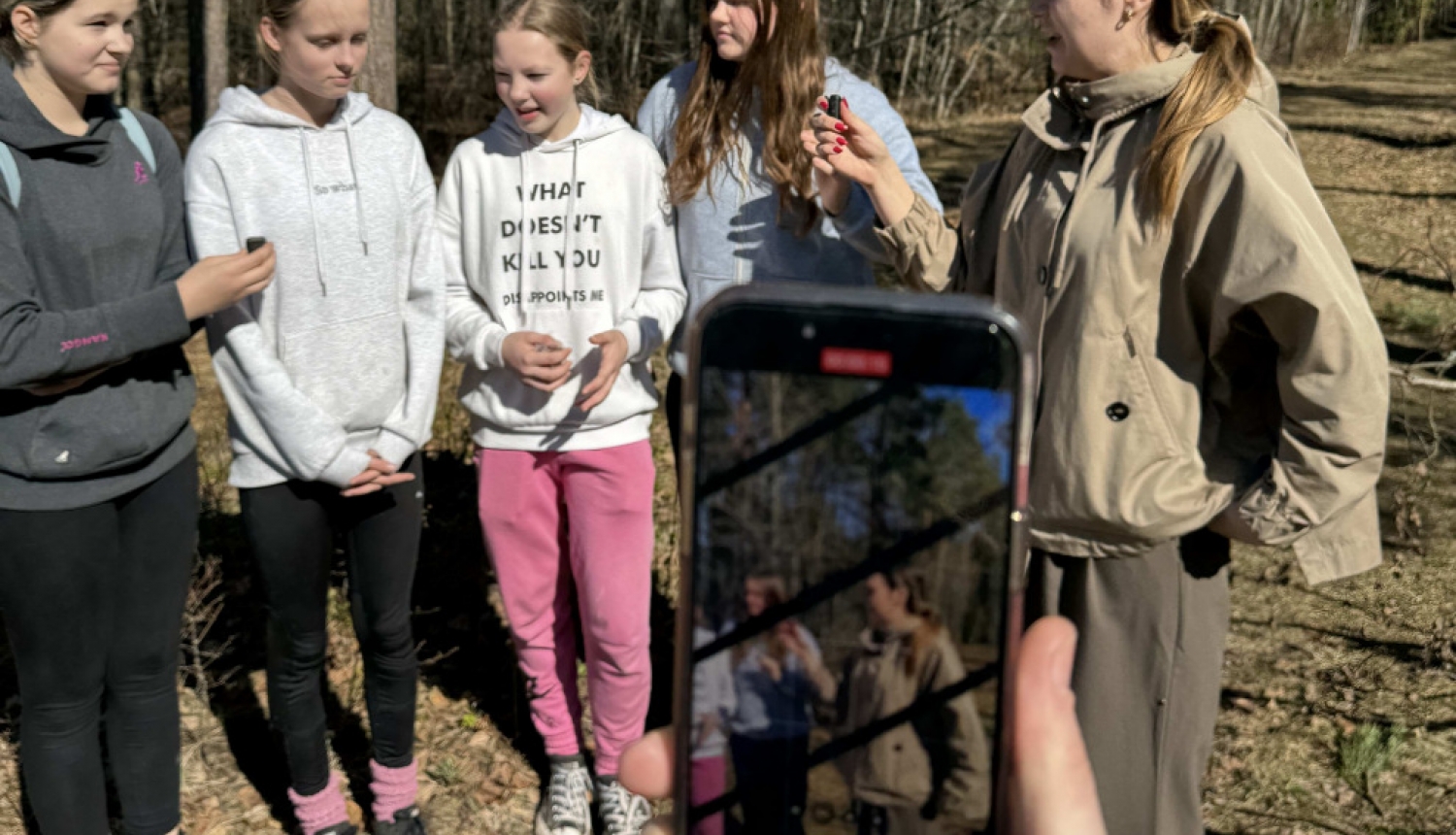 Piecas jaunietes stāv un sarunājas ārā, aizmugurē koki, priekšā kāds filmē šo sarunu tiešraidē