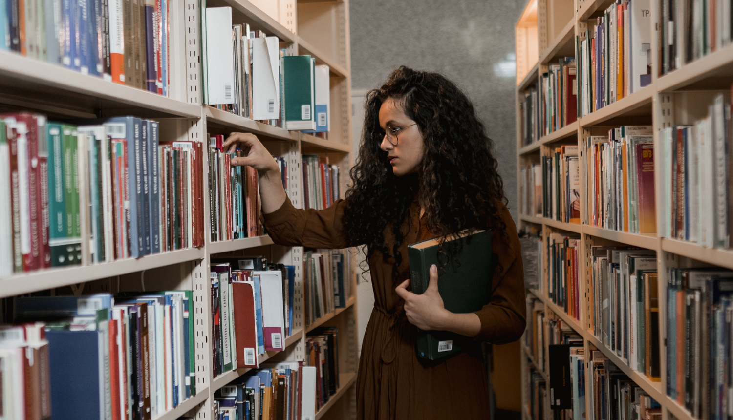 Tumšmataina sieviete  ar grāmatām rokās meklē grāmatas bibliotēkā grāmatu plauktā