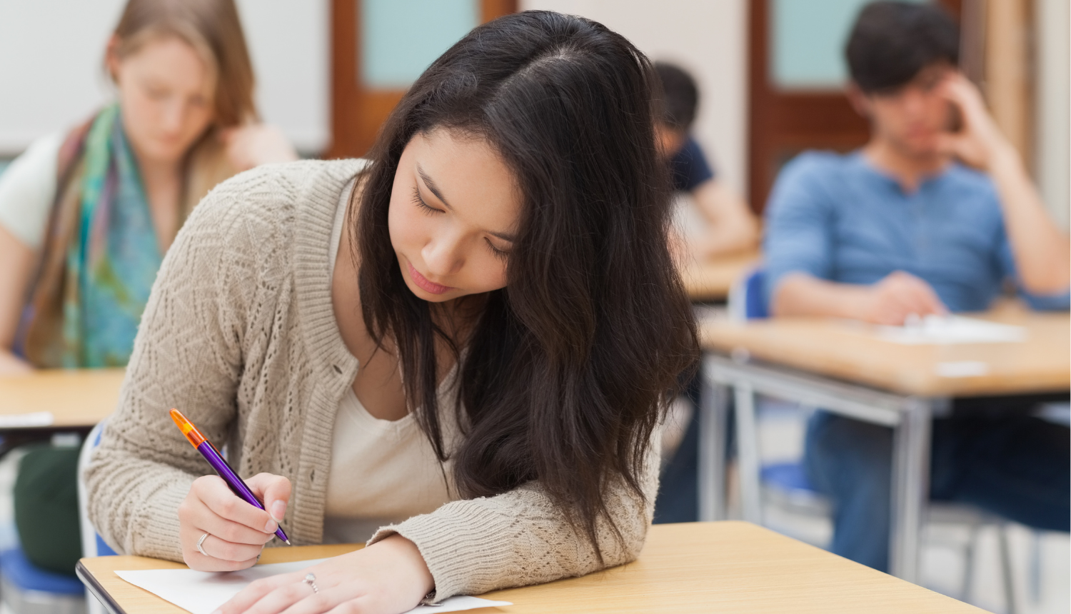 jaunieši klasē raksta eksāmenu, priekšplānā tumšmataina meitene sēž pie galda un raksta uz lapas, roku nolikusi uz lapas