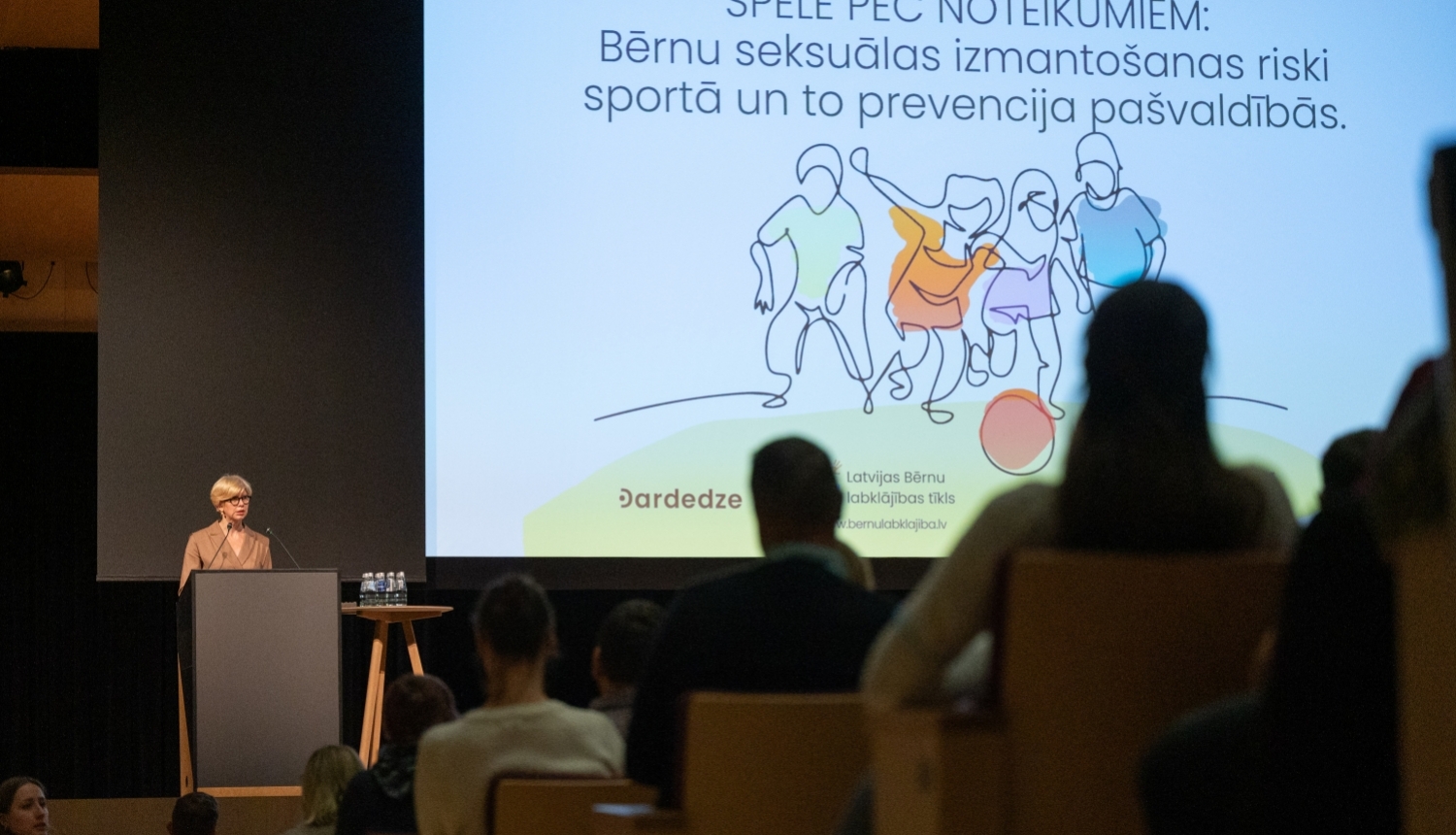 Izglītības un zinātnes ministre Anda Čakša konferencē uz skatuves, aiz viņas liels ekrāns ar bērnu ilustratīvu zīmējumu